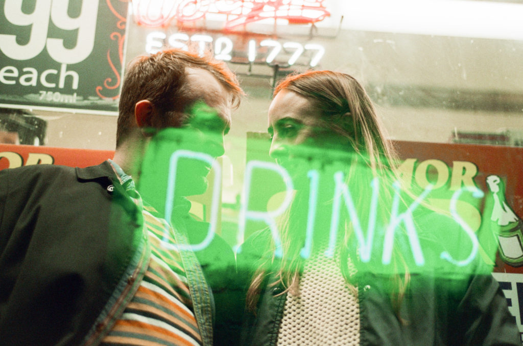 a couple looking at teachother with a neon sign that says drink